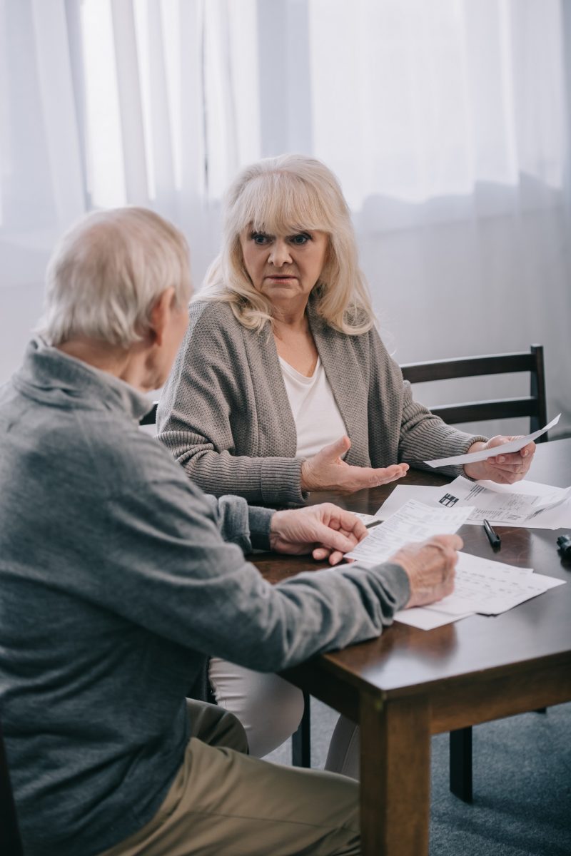 surprised senior couple in casual clothes sitting at table and holding bills